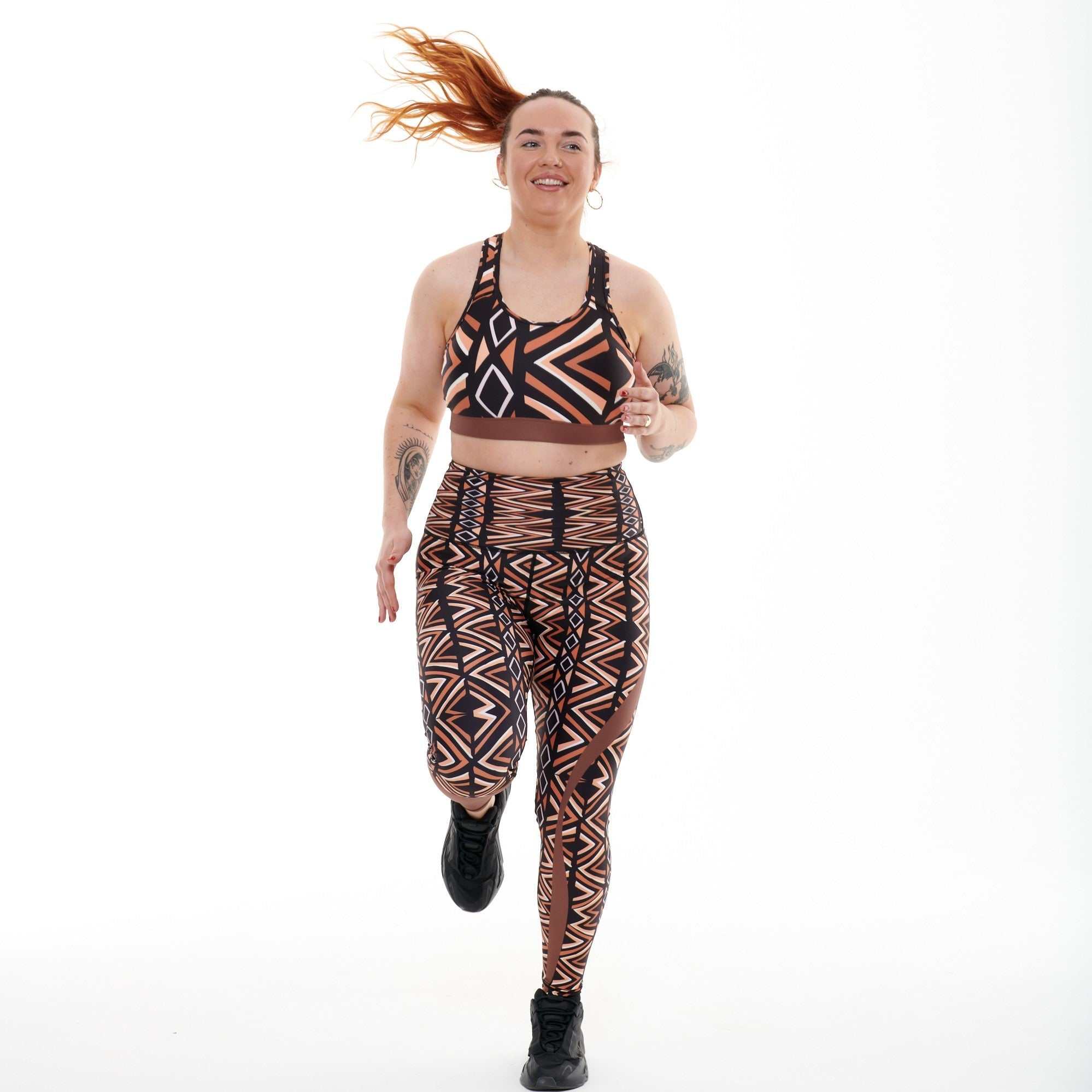 Vibrant Ibeekay leggings with Bògòlanfini Mud Cloth-inspired print in brown, orange, black, and white, featuring an engineered waistband and phone pocket.